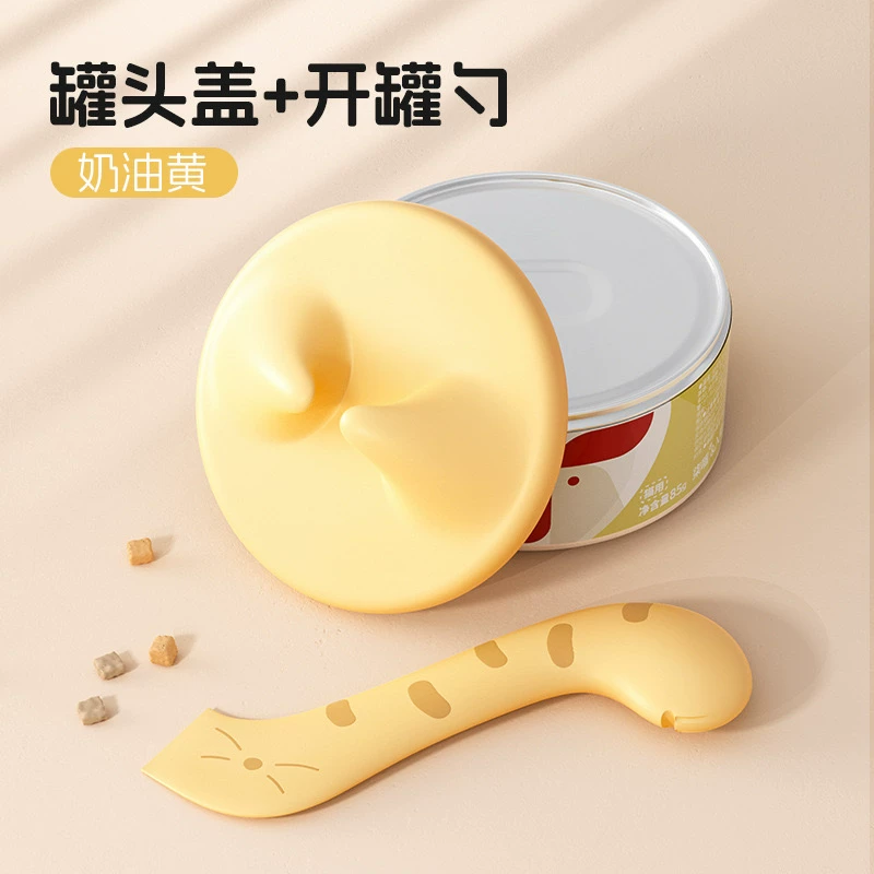 Supplier photo of the selected yellow can lid with two raised grips and curved striped cat-shaped spoon. One lid and one spoon supplied; can and food are display props.
